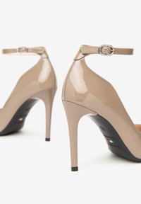 Béžové lakované kožené lodičky na vysokém podpatku s kotníkovými pásky a sponami posetými krystaly, zobrazené zezadu na bílém pozadí.