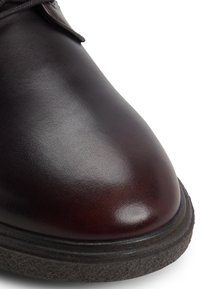Zapato de cuero marrón con un acabado brillante y suave. Presenta una puntera redondeada y una suela de goma negra texturizada. Los cordones son visibles sobre la lengua.