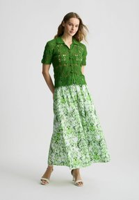 Top corto de ganchillo verde con patrón floral, combinado con una falda larga de color verde pastel con un diseño de hojas y tacones blancos de tiras.