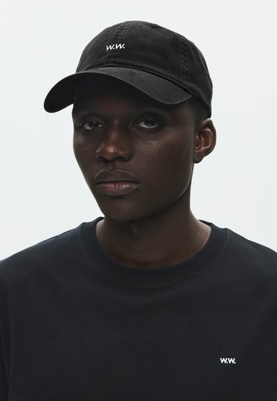 Fiatal férfi fekete baseballsapkában és hozzá illő fekete pulóverben, mindkét darabon kis fehér "W.W." logóval, semleges háttér előtt.