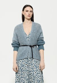 Hellblau gestrickte Strickjacke mit tiefem V-Ausschnitt, gerippten Bündchen und Taillengürtel, die über einem floral gemusterten Kleid in Blau- und Weiss-Tönen getragen wird.