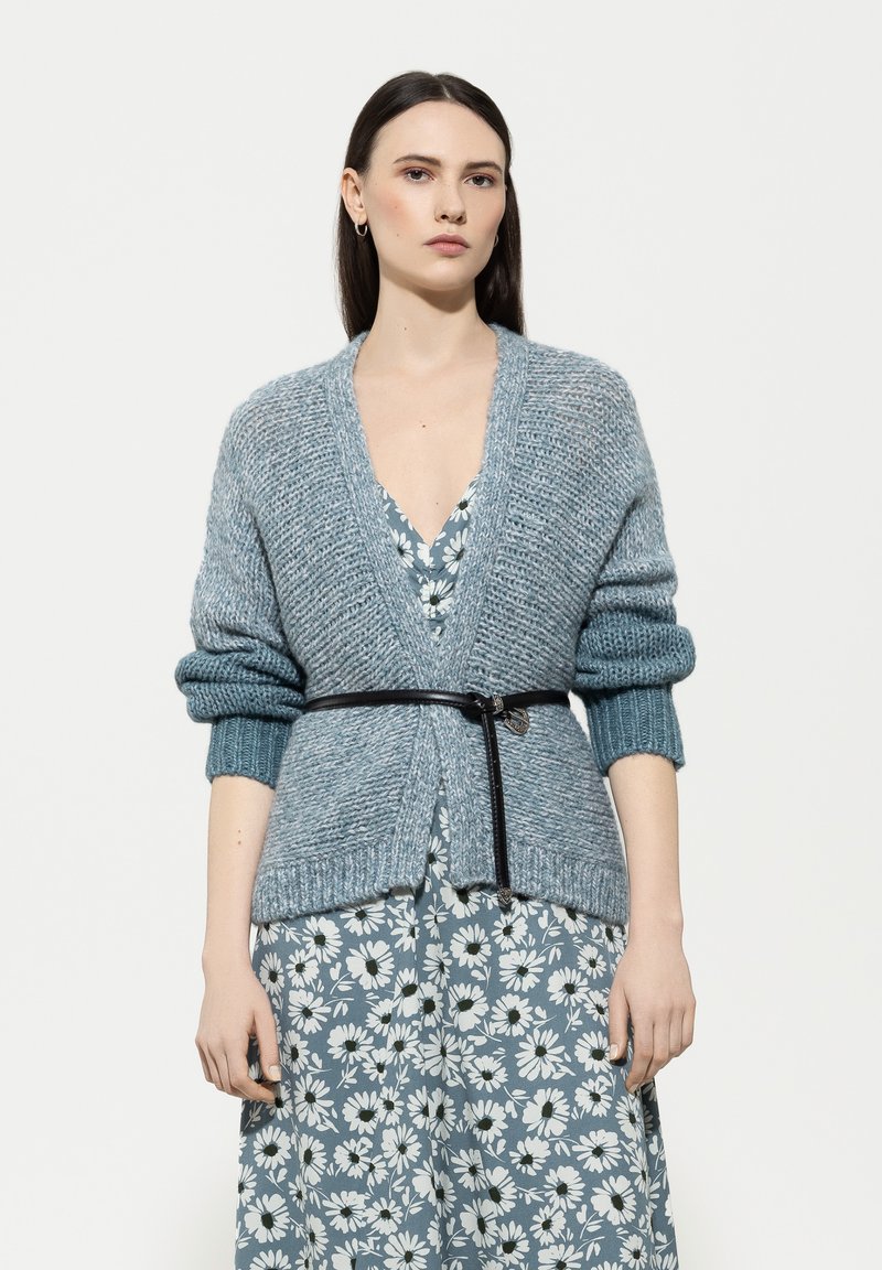 Hellblau gestrickte Strickjacke mit tiefem V-Ausschnitt, gerippten Bündchen und Taillengürtel, die über einem floral gemusterten Kleid in Blau- und Weiss-Tönen getragen wird.