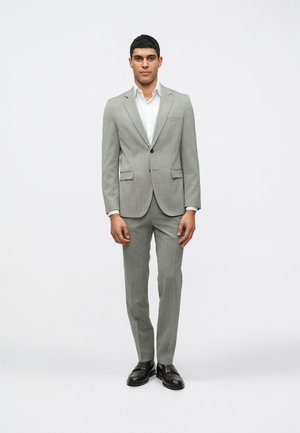 Šviesiai pilkas pritaikytas kostiumas su subtiliomis juostelėmis, blazeris su vienu mygtuku, suapvalintais atvartais ir įliemenuotomis kelnėmis. Po juo dėvima balta marška.