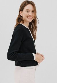 Czarny kardigan z jasnym wykończeniem, o prążkowanej strukturze i z detalem guzika. Długie rękawy i krótki krój, noszony na jasnych spodniach.