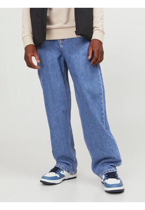 Personne portant un jean bleu ample, des baskets blanches et bleues, un haut beige à manches longues et un gilet noir, debout devant un fond uni.