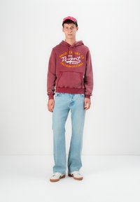 Sudadera burdeos con texto gráfico amarillo, corte relajado, bolsillo canguro; vaqueros de pierna ancha azul claro; zapatillas blancas con suelas marrones.