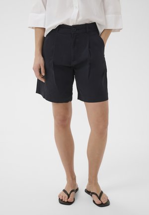 Femme portant un short noir boutonné, une chemise blanche aux manches retroussées et des tongs noires, debout devant un fond blanc uni.