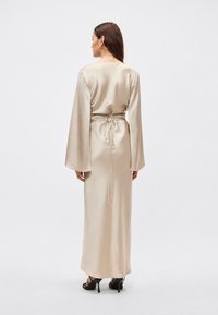 Ilga šviesiai smėlio spalvos satino suknelė su plačiomis varpelio formos rankovėmis, žemu nugaros iškirpimu ir surišamu juosmens detaliu, vilkima su juodais bateliais su dirželiais.