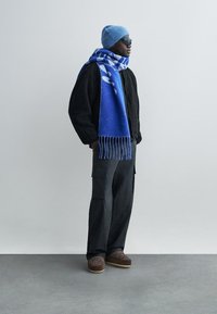 Zwarte trui met textuur, blauw sjaal met patroon, lichtblauwe gebreide muts, donkere cargobroek en bruin patroon schoeisel. Staand tegen een effen muur.