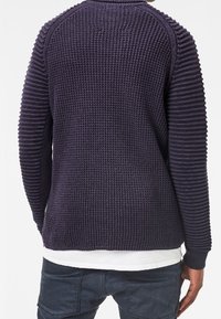 Mörklila stickad tröja med strukturerad ribbning på ärmar och kropp, med rund halsringning och avslappnad passform.