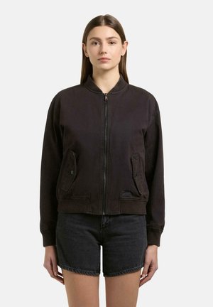 Donna con capelli castani lisci che indossa una giacca bomber nera con cerniera e shorts di denim neri, in piedi davanti a uno sfondo semplice.