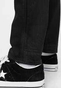 Musta teksturoitu farkkuhousut, jotka on yhdistetty mustiin mokkanahkakenkiin, joissa on valkoinen tähden logo, valkoiset pohjat ja erottuvat valkoiset nauhat.