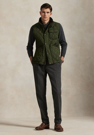 Hombre con chaleco acolchado verde oscuro sobre suéter gris oscuro, pantalones grises y zapatos de cuero marrón, de pie con las manos en los bolsillos.