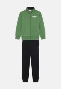 Sudadera verde con cremallera y cuello alto, combinada con pantalones de chándal negros con cordón y un pequeño logo.
