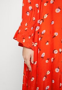 Χέρι να ξεκουράζεται δίπλα σε ένα κόκκινο φόρεμα με ροζ και λευκή λουλούδια εκτύπωση, που διαθέτει μαζεμένα ελαστικά μανίκια στις μακριές μανσέτες.