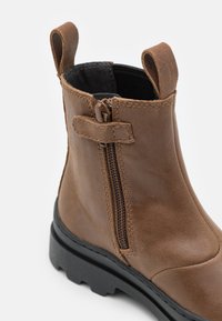 Bottine en cuir brun avec une semelle en caoutchouc noir, fermeture éclair latérale et anses pour un enfilage facile. Texture lisse avec des détails de couture subtils.