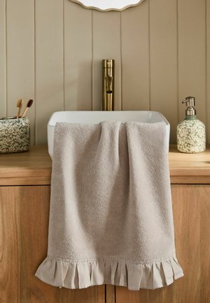 Beige håndklæde med flagrende kant hængende over et træskab under en hvid vask med guldhane, blomster tandbørsteholder og sæbedispenser.