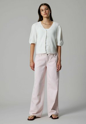 Mujer joven de pie, con cárdigan blanco de cuello en V, pantalones anchos de color rosa claro, collar dorado y sandalias sobre un fondo gris liso.