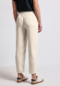 Light beige linen trousers with an elastic waistband, relaxed fit, and two back pockets. Paired with white and beige sneakers.