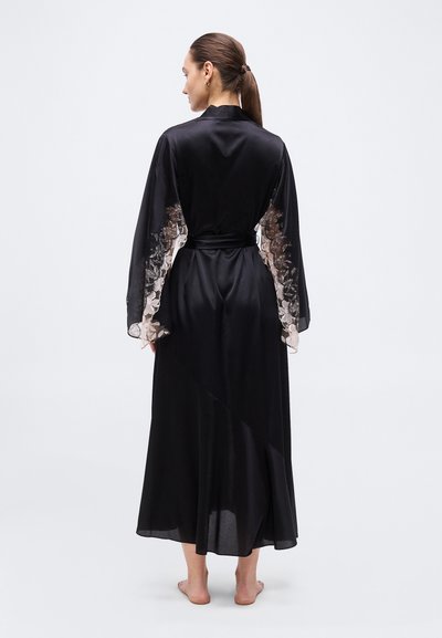 Γυναίκα που στέκεται ξυπόλητη, φορώντας μακρύ μαύρο σατέν ρόμπα με διαφανείς μανίκια από δαντέλα με floral σχέδιο, φωτογραφημένη από πίσω πάνω σε απλό ανοιχτό φόντο.