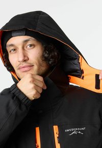 Zwarte waterdichte jas met een oranje binnenkant, voorzien van een hoge kraag, elastische manchetboorden en een logo op de borst.