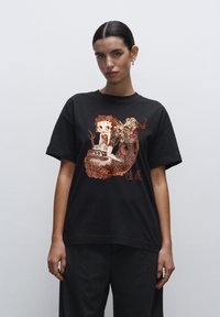 Camiseta negra de algodón, con un gráfico divertido en tonos rojos apagados y crema de un personaje con un fondo de dragón.