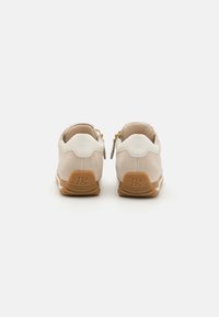 Beige semskinn sneakers med hvite lærdetaljer og gullglidelås, med en tan gummisåle og logo på baksiden. Flat hældesign.