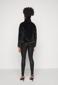 Chaqueta de piel sintética negra con un patrón en zigzag, cuello alto y mangas largas, combinada con pantalones de cuero negros y tacones con tiras.