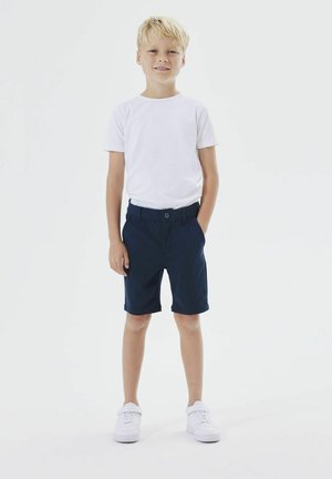 Maglietta bianca a maniche corte, pantaloni corti blu navy con passanti per cintura e tasche anteriori, e sneakers bianche, su un ragazzo in piedi con le mani nelle tasche.