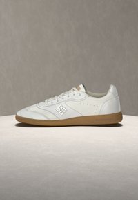 Λευκό sneaker χαμηλού τύπου με λαστιχένια σόλα, λογότυπο στο πλάι και κορδόνια, τοποθετημένο σε φωτεινή επιφάνεια σε ουδέτερο φόντο.