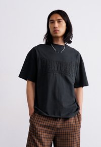 Black oversized t-shirt with textured logo on the front, paired with brown plaid trousers. Simple design, crew neckline, short sleeves.