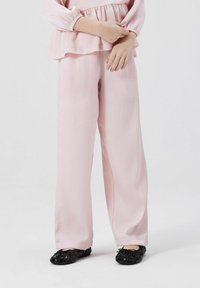 Helle rosa, strukturierte Bluse mit langen Ärmeln und Rüschen, kombiniert mit locker sitzenden, hellrosa Hosen. Schwarze strukturierte Ballerinas runden das Outfit ab.