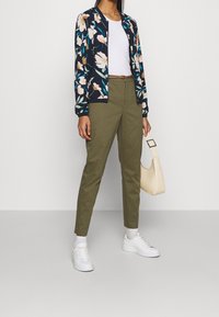 Kvinna i olivgröna byxor, vita sneakers, vit t-shirt, blommig zip-up jacka, som håller en beige handväska framför en enfärgad bakgrund.