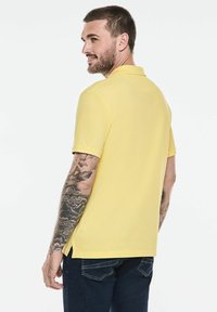 Žluté polo tričko s krátkými rukávy, bočními rozparky a klasickým límečkem, kombinované s tmavě modrými džíny. Viditelné kožní tetování na pažích.
