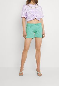 T-shirt cropped en tie-dye violet clair avec des manches retroussées, associée à un short en denim vert menthe avec une fermeture à boutons et des poches avant.