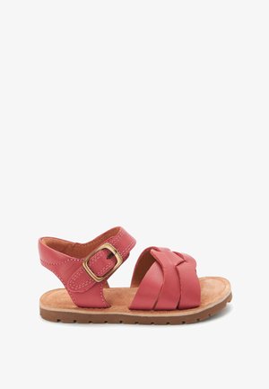 Lyserød lædersandal med crossover-remdesign, justerbar spænde og brun ruskindsål. Har en flad gummisål med teksturerede detaljer.