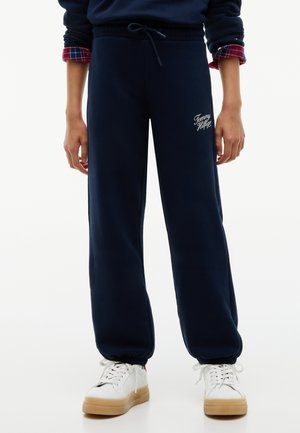 Person iført marineblå Tommy Hilfiger joggingbukser med elastiske manchetter og hvide sneakers med gummisåler.