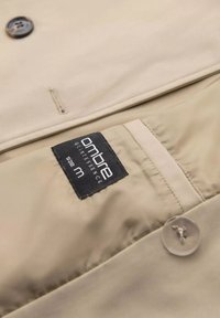 Beżowy materiał kurtki o gładkiej fakturze, z plastikowym guzikiem oraz czarną etykietą z napisem "ombre quintessence rozmiar m."