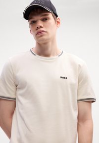 T-shirt crème avec col et poignets à rayures noires, arborant un petit logo "BOSS" noir sur le devant, en tissu texturé. Casquette noire avec logo.
