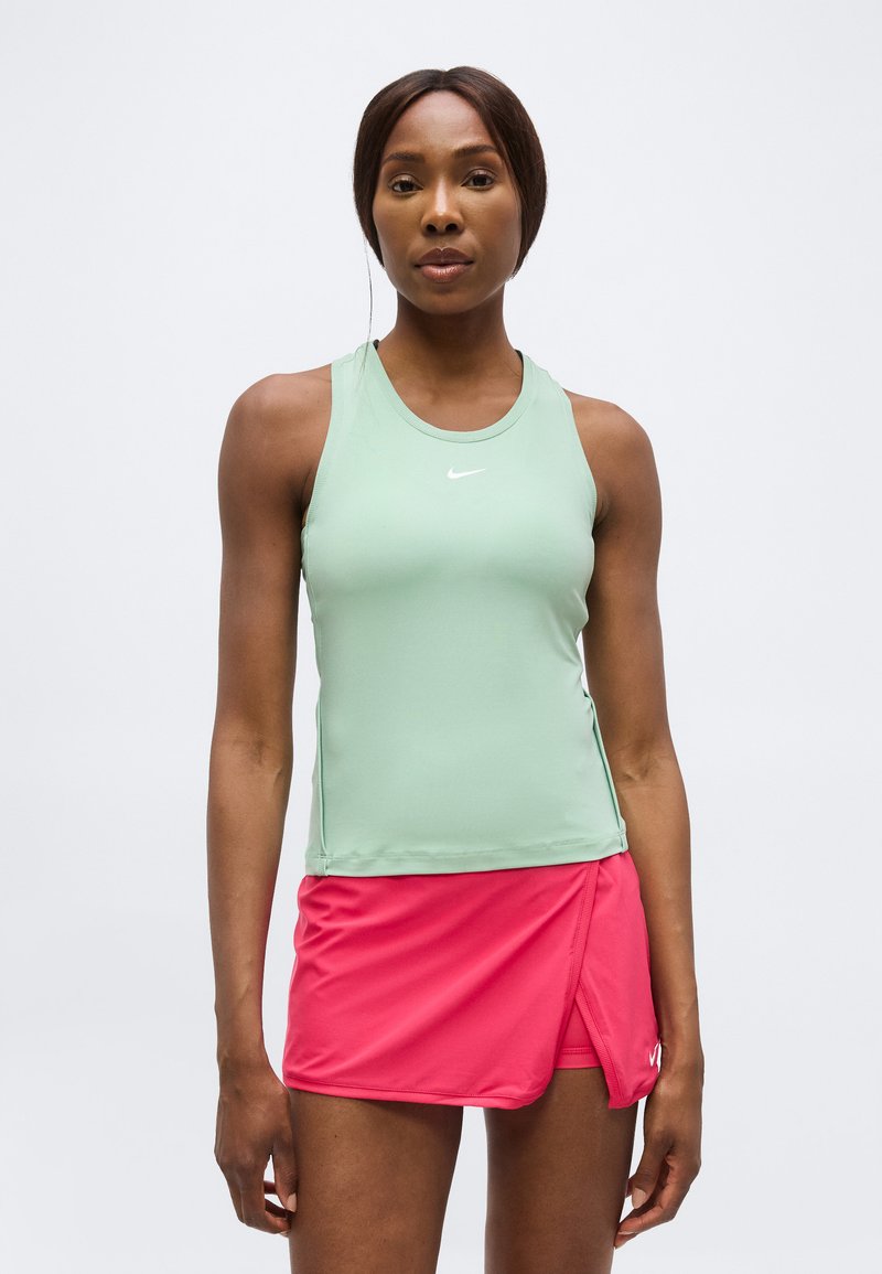 Femme debout portant un haut sans manches vert clair et une jupe-culotte de sport rose vif sur un fond blanc uni.