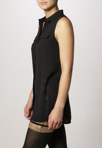 Kvinna klädd i en ärmlös svart skjorta med knappar och krage, genomskinliga svarta strumpbyxor och en paljetterad guldfärgad strumpeband.