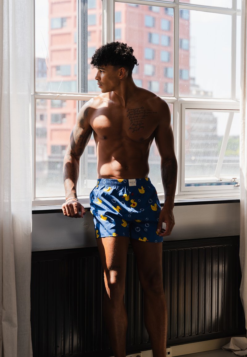 Pantaloni corti blu con un motivo di anatre di gomma, dotati di una cintura elastica. Il modello si trova accanto a una finestra, mettendo in risalto il design giocoso dei pantaloni.