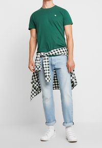 Grünes Baumwoll-T-Shirt mit kleinem Logo auf der Brust, kombiniert mit hellblauen Jeans. Ein schwarz-weiß karierter Flanellstoff um die Taille gebunden. Weiße Sneakers.