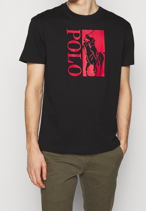 Mężczyzna w czarnej koszulce z czerwonym logo Polo i grafiką gracza polo, w połączeniu z oliwkowymi spodniami.
