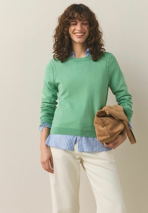 Vrouw met krullend haar die lacht, draagt een groene trui over een blauw gestreept overhemd, witte broek en houdt een beige suède tas vast.