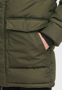 Tamnozelena jakna od puf materijala sa prešanom teksturom, bočnim džepom na zip, elastičnim manžetama, te oznakom brenda na donjem šavu.