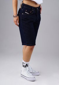Woman wearing dark denim knee-length shorts, white high-top sneakers, white socks with black text, and bracelets on her left wrist.