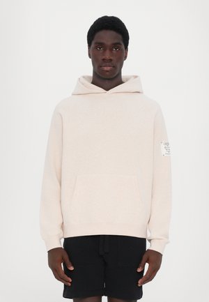 Pullover hoodie in light beige fabric with a kangaroo pocket, ribbed cuffs, and a label on the right sleeve; paired with black shorts.