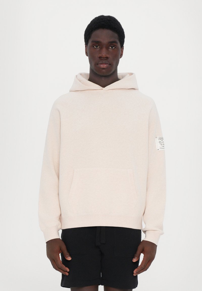 Pullover hoodie in light beige fabric with a kangaroo pocket, ribbed cuffs, and a label on the right sleeve; paired with black shorts.
