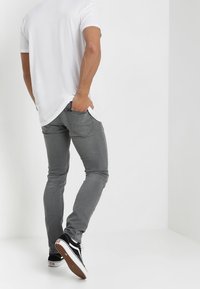 Šedé slim-fit džíny s zadními kapsami, kombinované s bílým tričkem a černými teniskami, zobrazené ze zadního úhlu na jednobarevném pozadí.